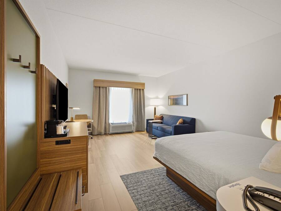 Habitación de hotel con cama, escritorio, silla, televisión, lámpara y ventana con cortinas.