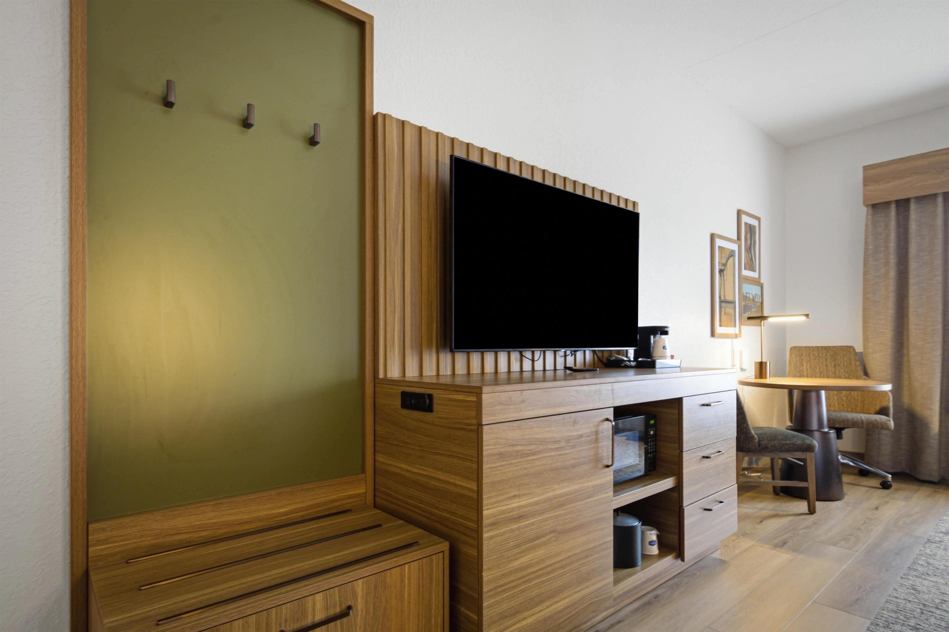 Habitación de hotel con una televisión de pantalla plana montada en un mueble de madera, un escritorio con lámpara, una silla y una cama con cabecero de madera.