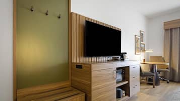 Habitación de hotel con una televisión de pantalla plana montada en un mueble de madera, un escritorio con lámpara, una silla y una cama con cabecero de madera.
