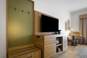 Habitación de hotel con una televisión de pantalla plana montada en un mueble de madera, un escritorio con lámpara, una silla y una cama con cabecero de madera.