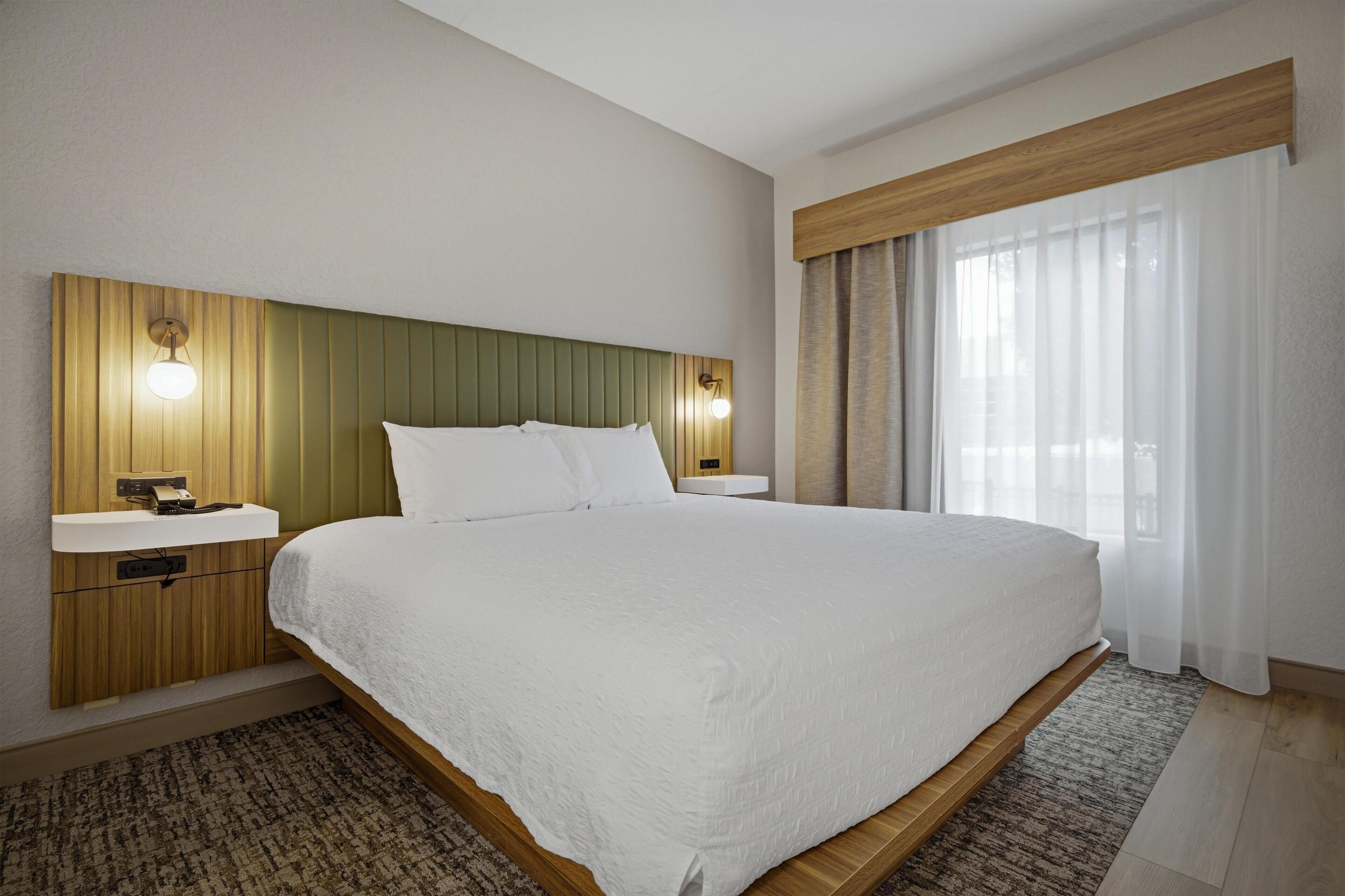 A hotel room with a large bed, wooden headboard, bedside lamps, a bedside telephone, and a window with curtains.