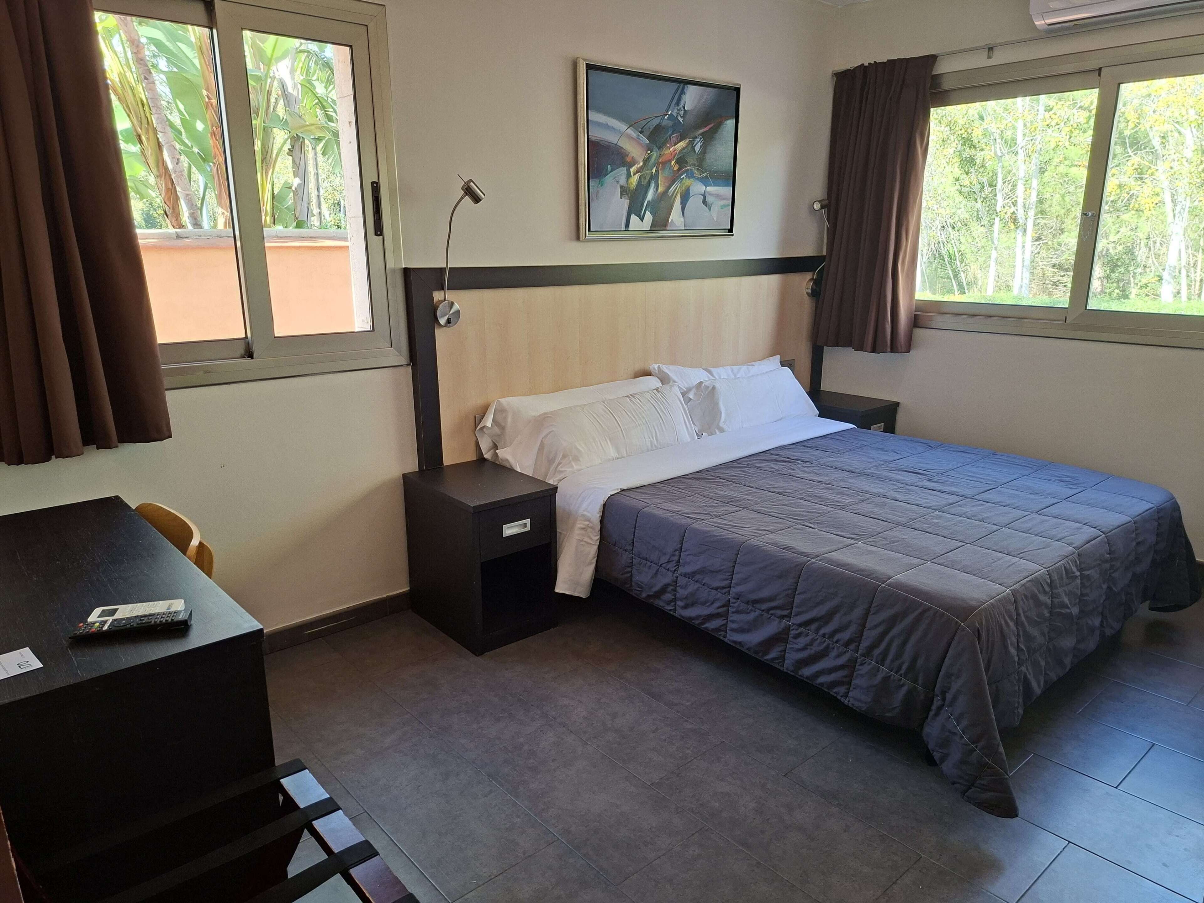 A hotel room with a bed, a desk with a remote control, a nightstand, and a painting on the wall.