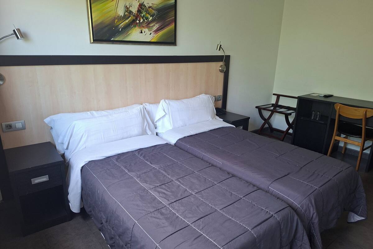 Una habitación de hotel con cama, escritorio, silla, mesita de noche y un cuadro en la pared.