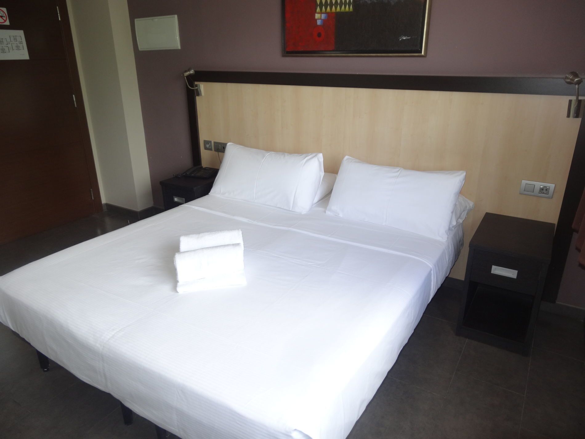 A hotel room with a large bed, two bedside tables, a nightstand, and a wall-mounted artwork.