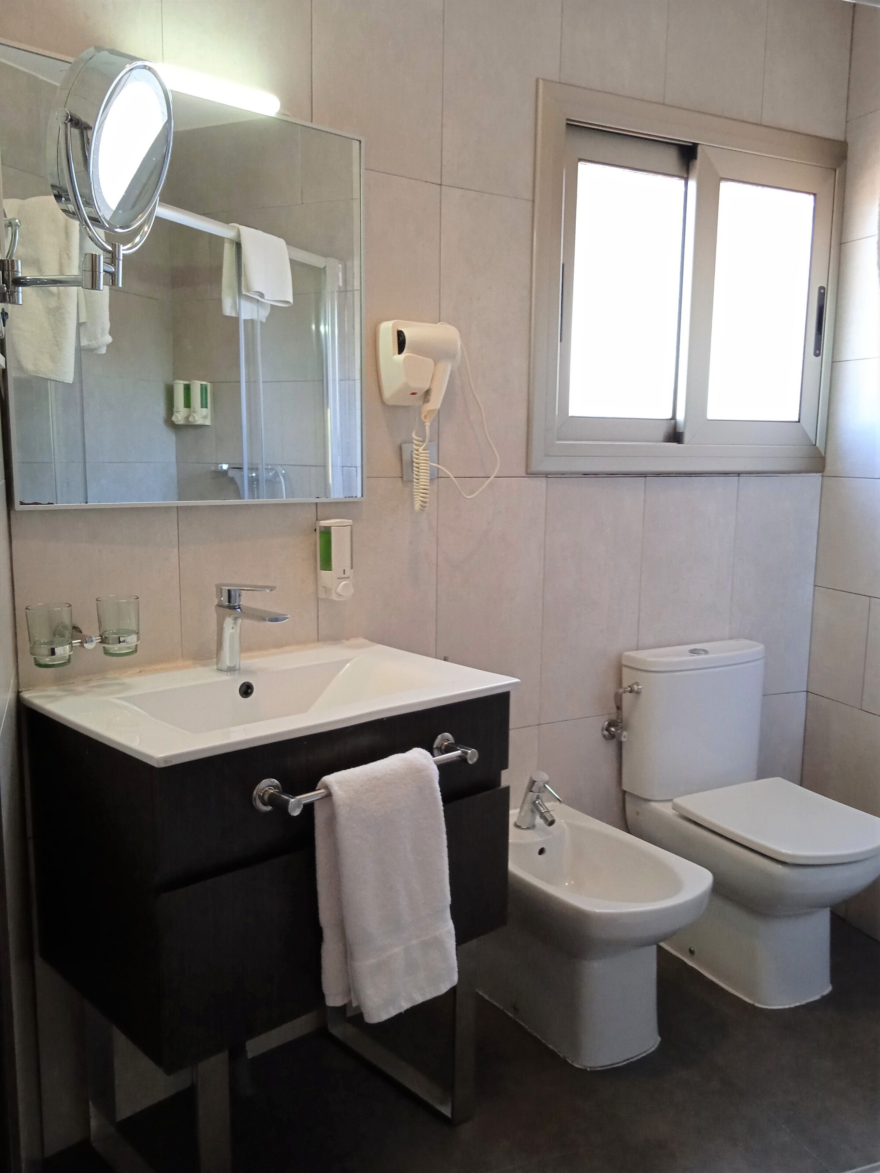 Une salle de bain équipée d’un lavabo, d’un miroir, d’un porte-serviettes, de toilettes, d’un bidet et d’un sèche-cheveux fixé au mur.