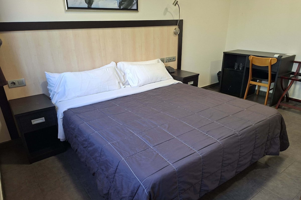 Una cama bien hecha con una colcha oscura, una mesita de noche con una lámpara y un escritorio con una silla.