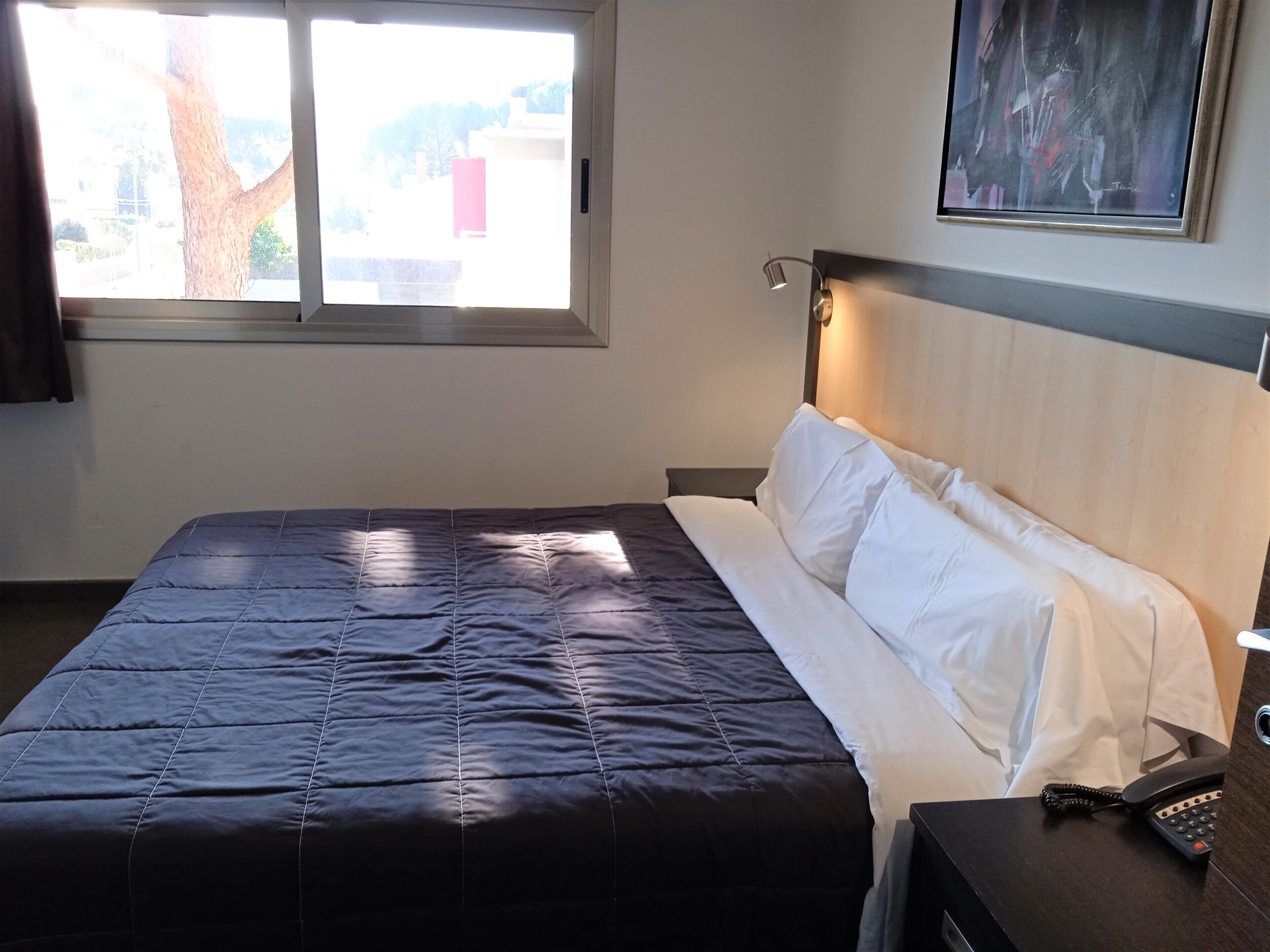 Une chambre d’hôtel avec un lit, une table de chevet avec un téléphone, un tableau au mur et une fenêtre donnant sur des immeubles.