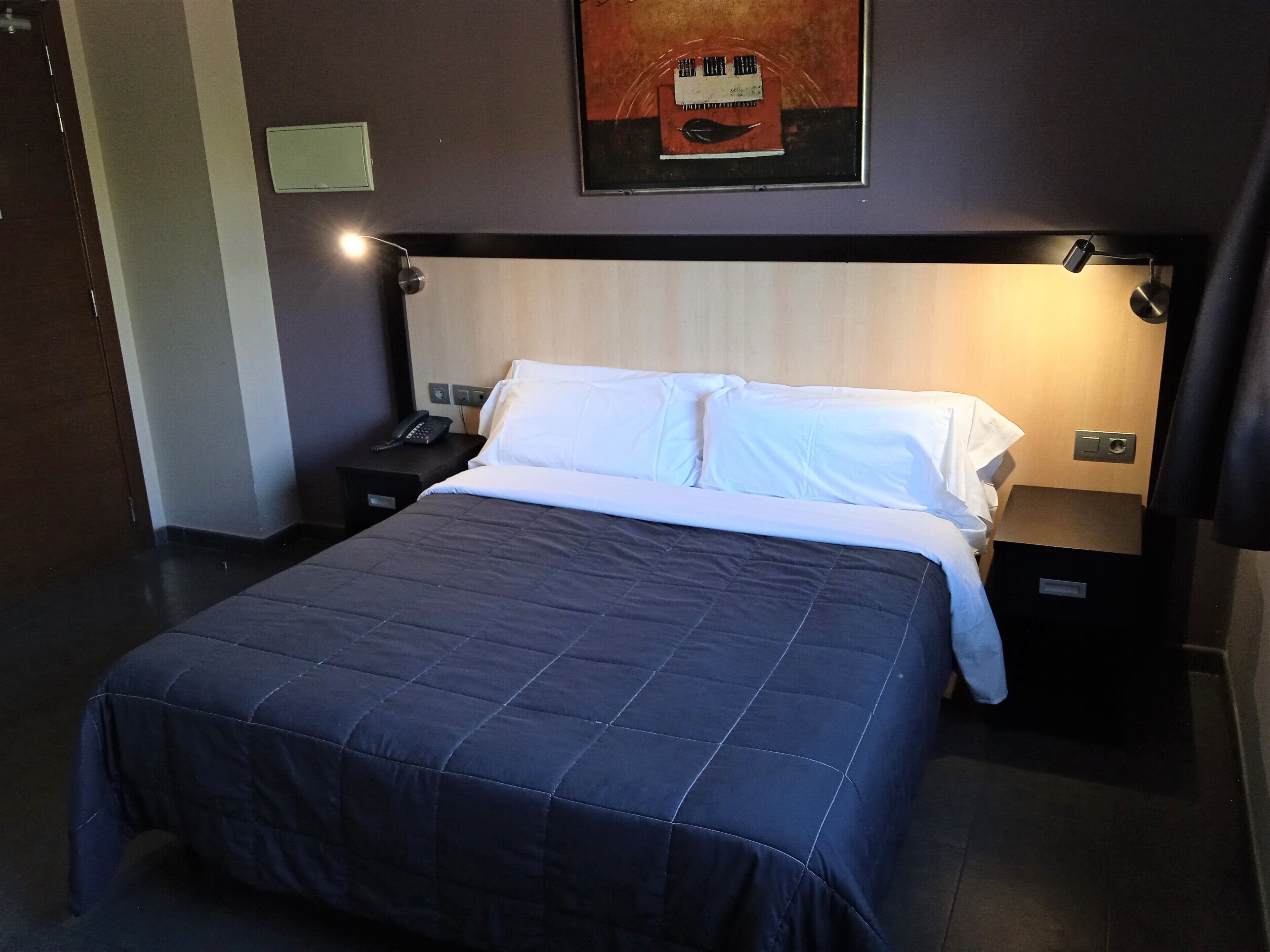 A hotel room with a bed, bedside tables, a phone, and a wall-mounted light fixture.