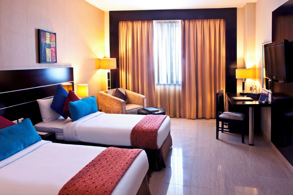 Habitación de hotel con dos camas, un escritorio, una silla, un televisor y un ventanal con cortinas.