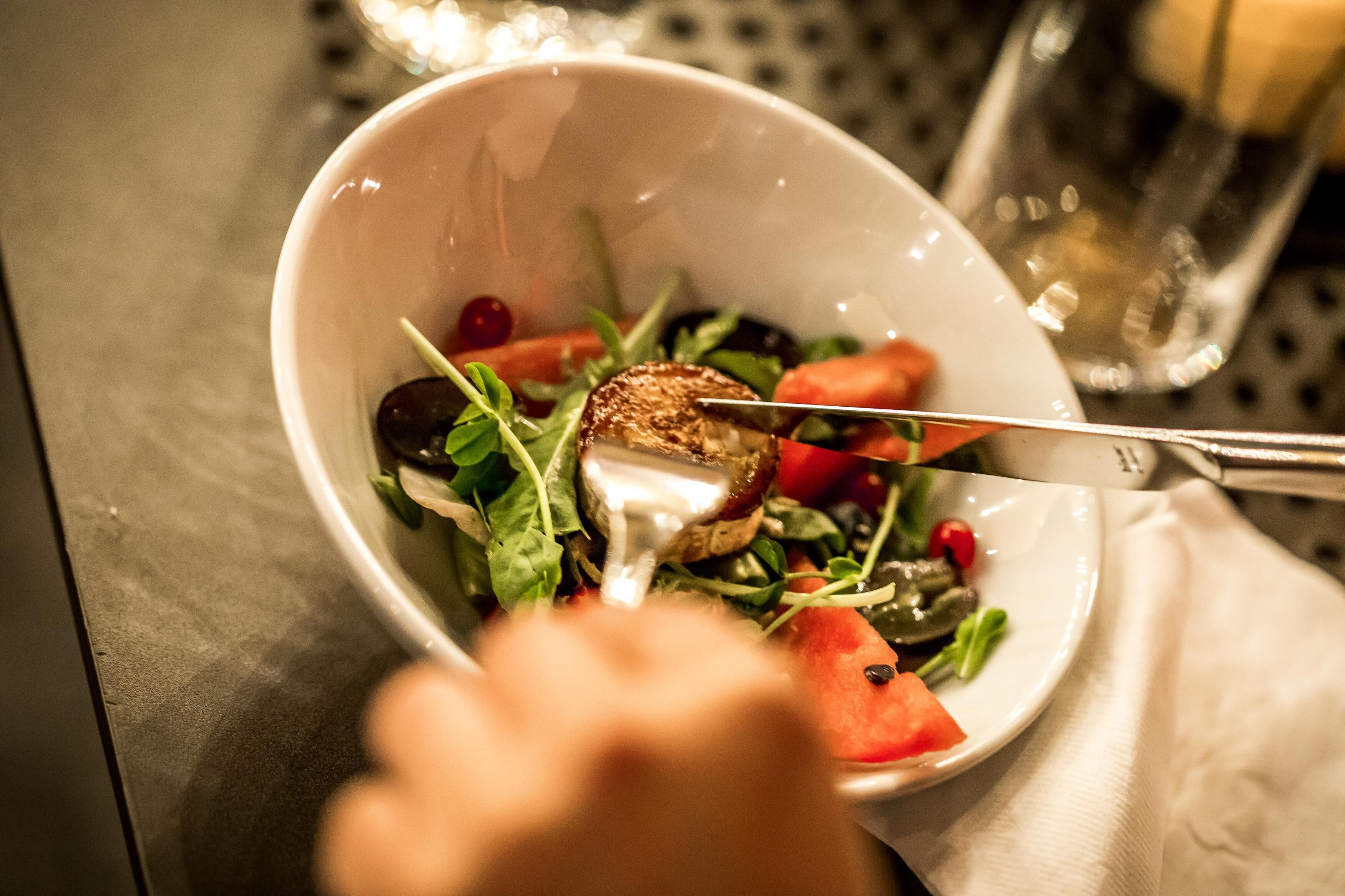 A white bowl containing a salad with greens, tomatoes, and a grilled item, possibly a meat or vegetable, on a table with a napkin and a glass.