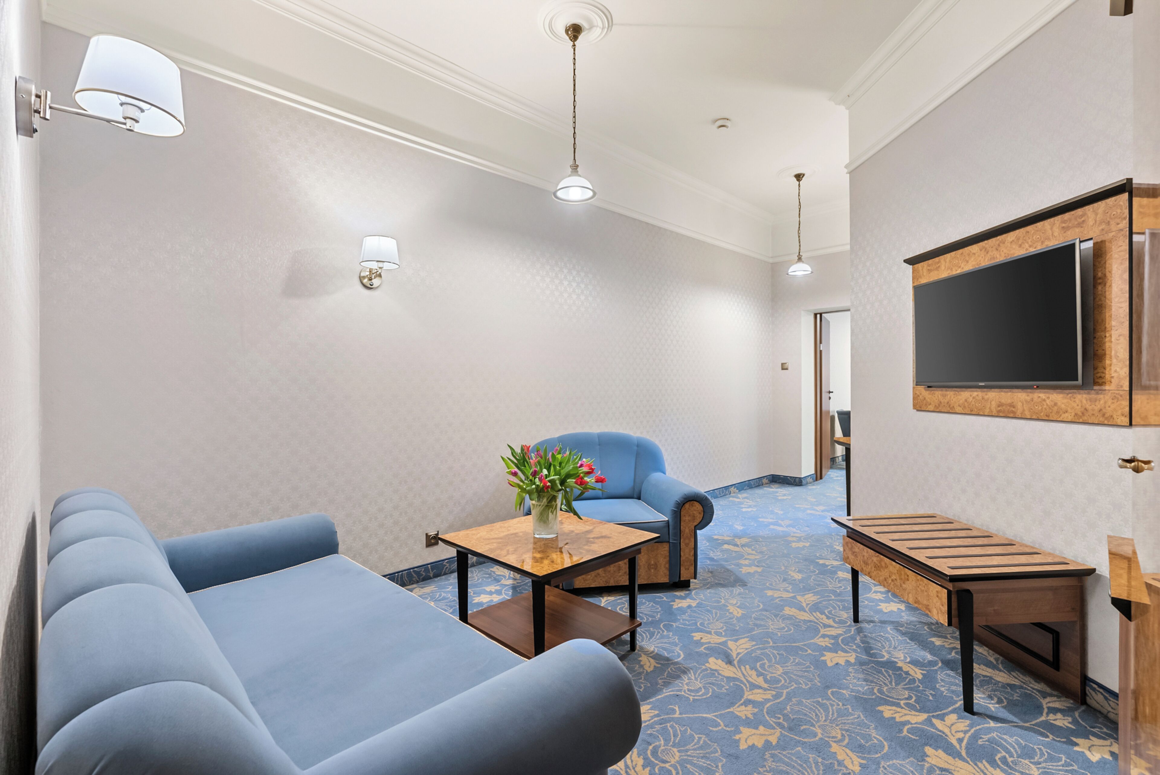 Una habitación de hotel con un sofá azul, una pequeña mesa con un jarrón de flores, un sillón azul, un banco de madera y un televisor de pantalla plana montado en la pared.