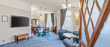 Habitación de hotel con una televisión de pantalla plana grande, un sofá, un comedor con mesa y sillas, y una alfombra estampada.