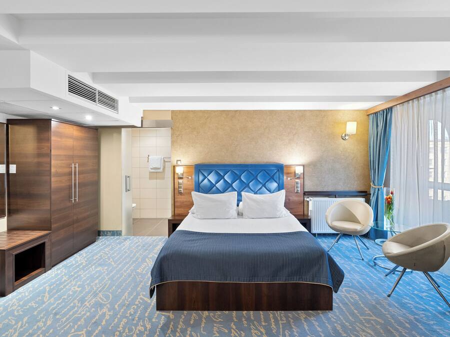 Habitación de hotel con una cama grande, zona de estar con una silla, escritorio y armario.