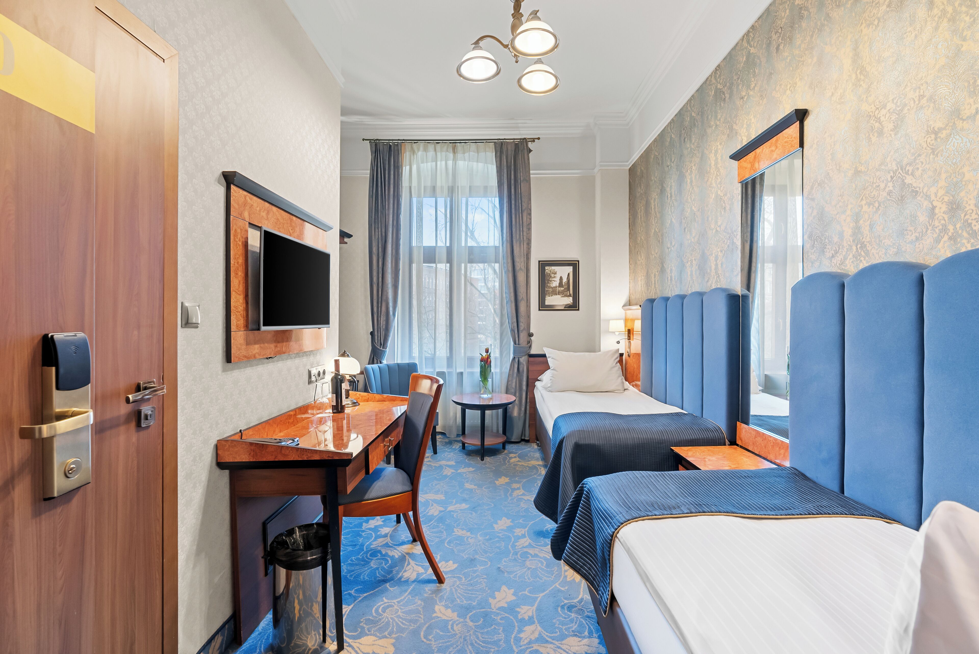 A hotel room with a desk, two beds, a TV, and a window with curtains.