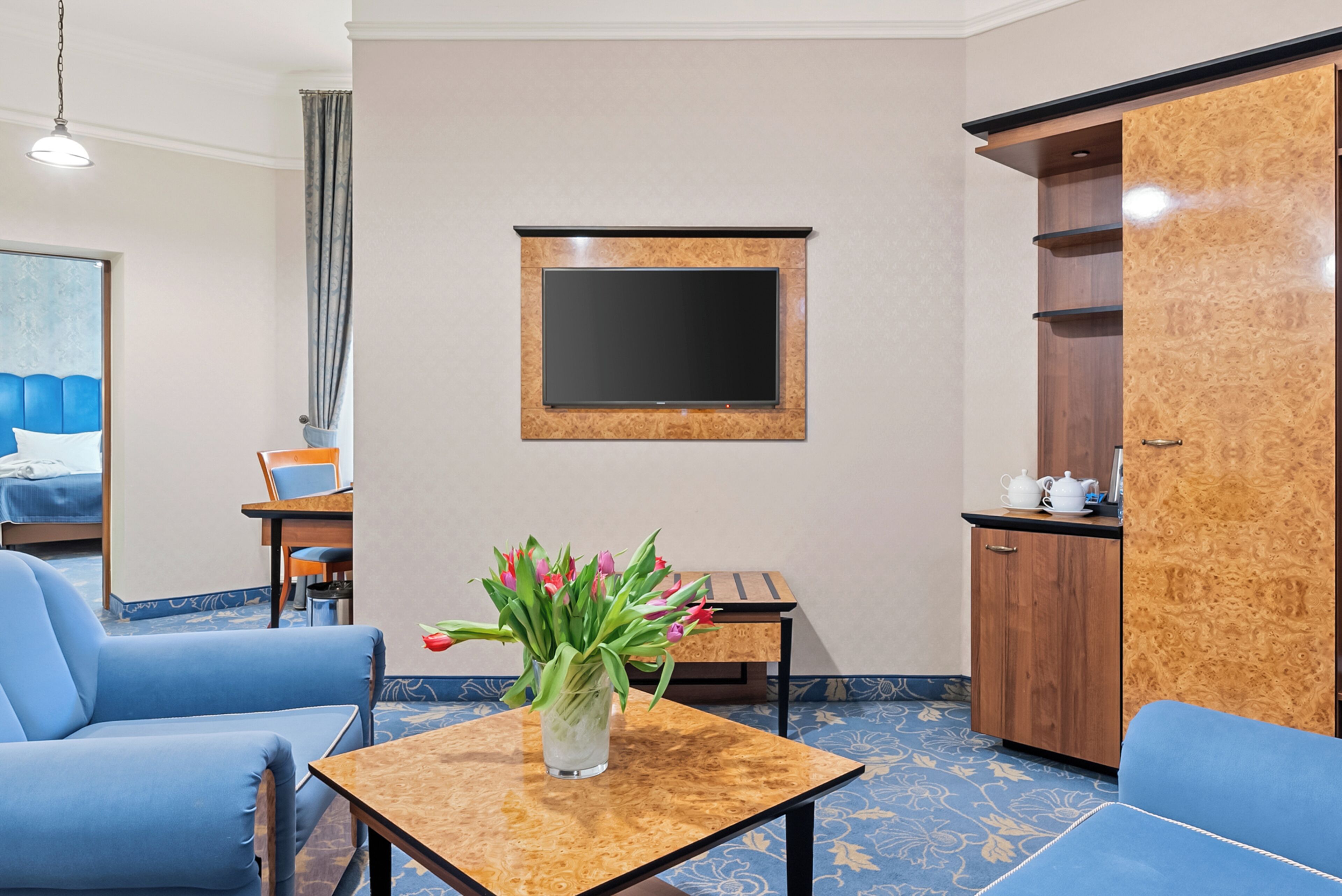 A hotel room with a bed, a blue sofa, a small table with a vase of flowers, a TV mounted on the wall, and a wooden cabinet.