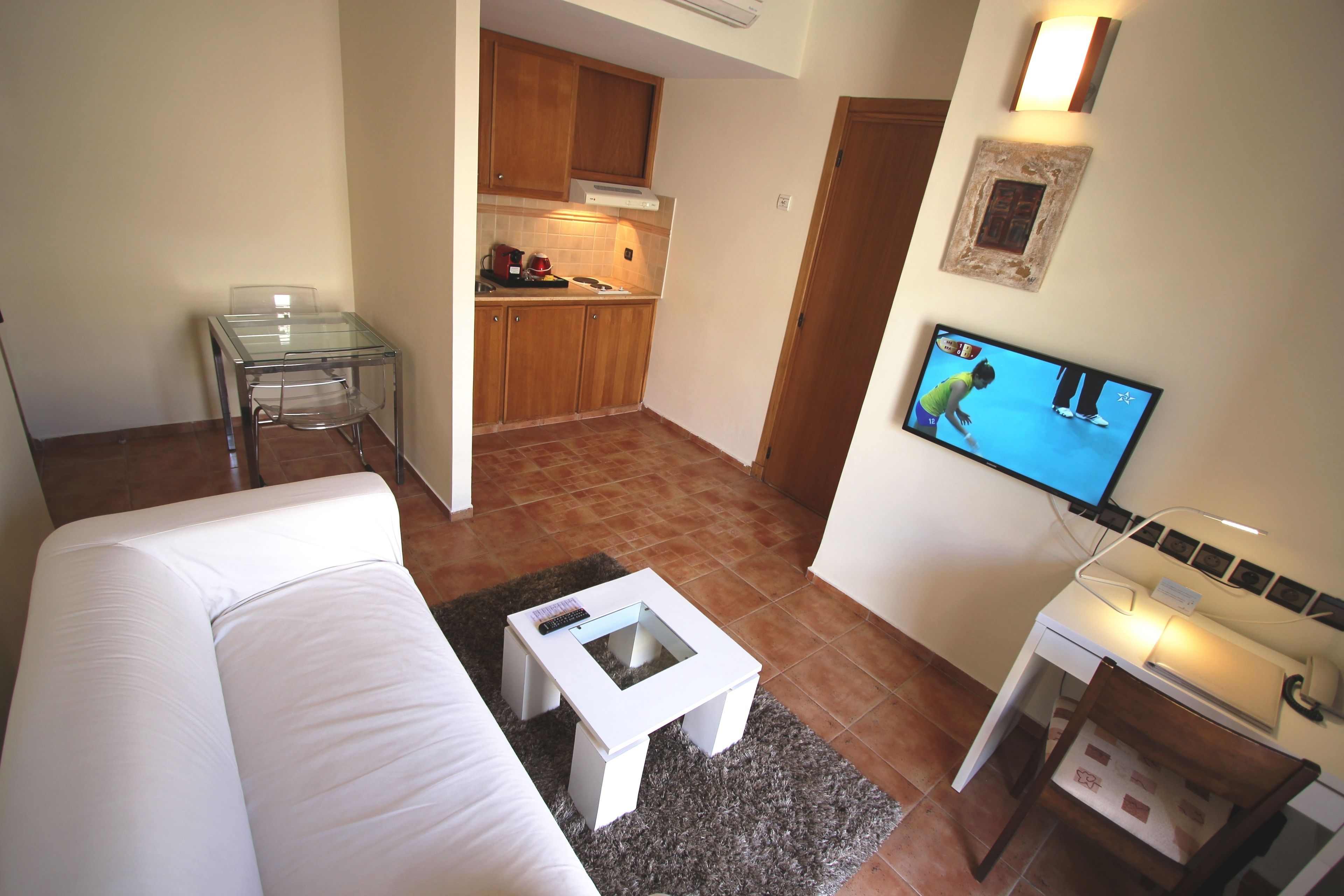 Una sala de estar con un sofá blanco, una mesa de centro de vidrio, un televisor y una zona de cocina con armarios de madera y un salpicadero de azulejos.