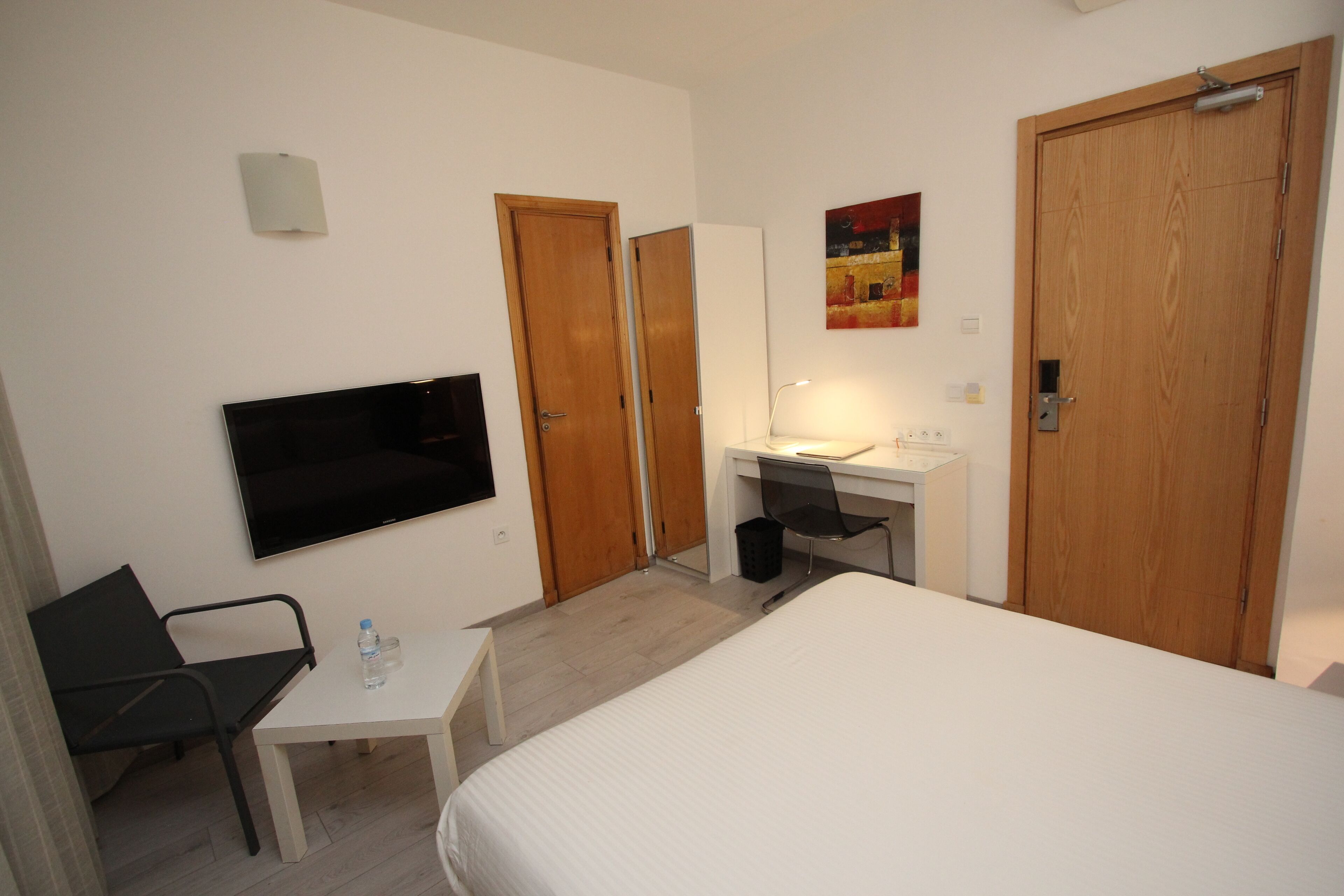 A hotel room with a bed, a desk, a chair, a TV, a mirror, and two doors.