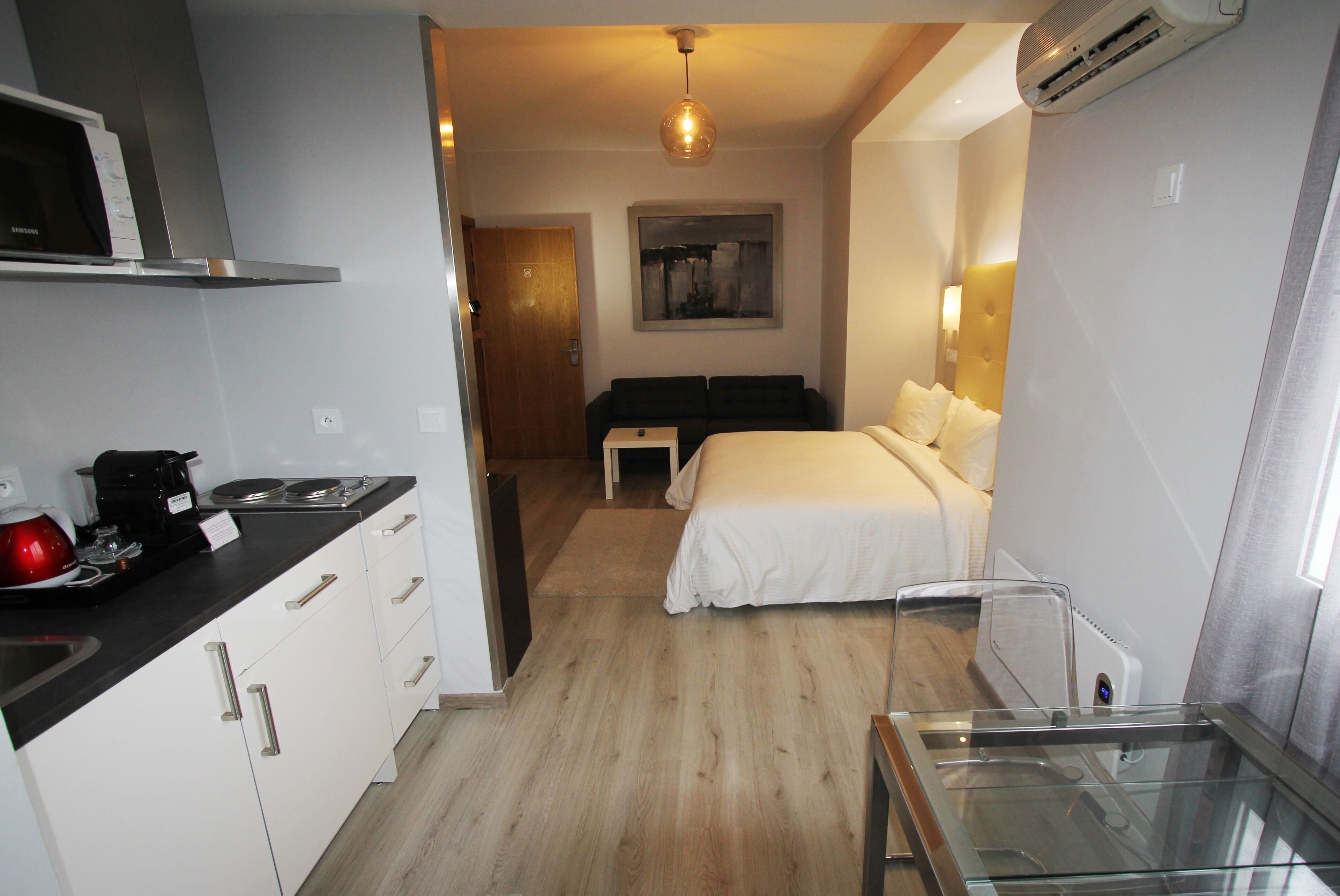 Una camera d'albergo compatta con angolo cottura, un letto con biancheria bianca, un piccolo comodino e un condizionatore d'aria a parete.