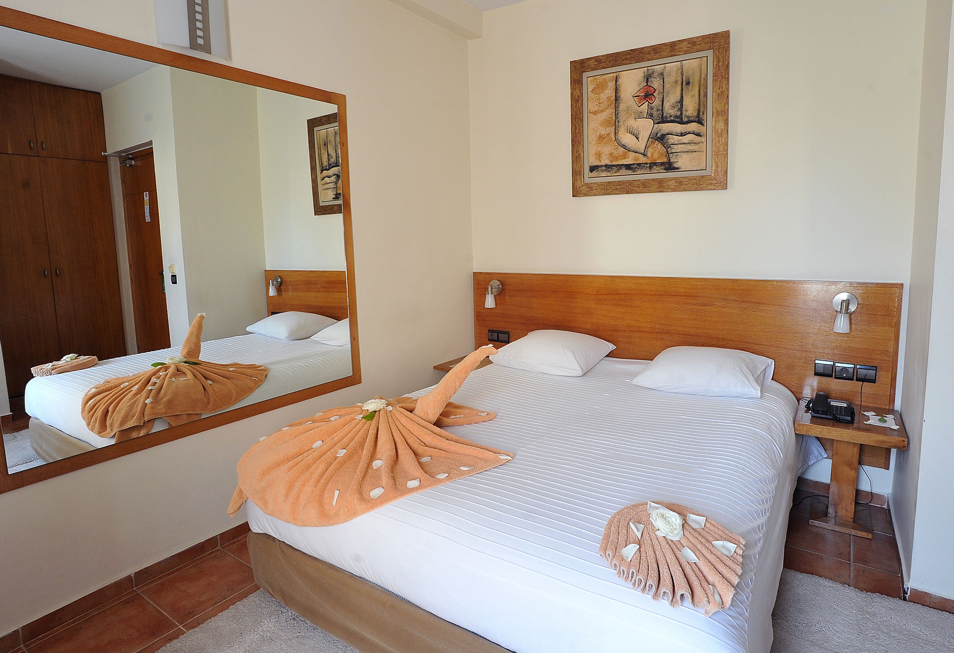 Quarto de hotel com uma cama grande, cabeceira de madeira, mesa de cabeceira com abajur, espelho e um quadro na parede.