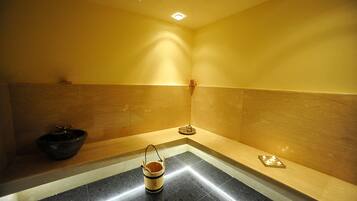 Una habitación pequeña y tenuemente iluminada con una bañera negra, un balde de madera y una bandeja con velas.