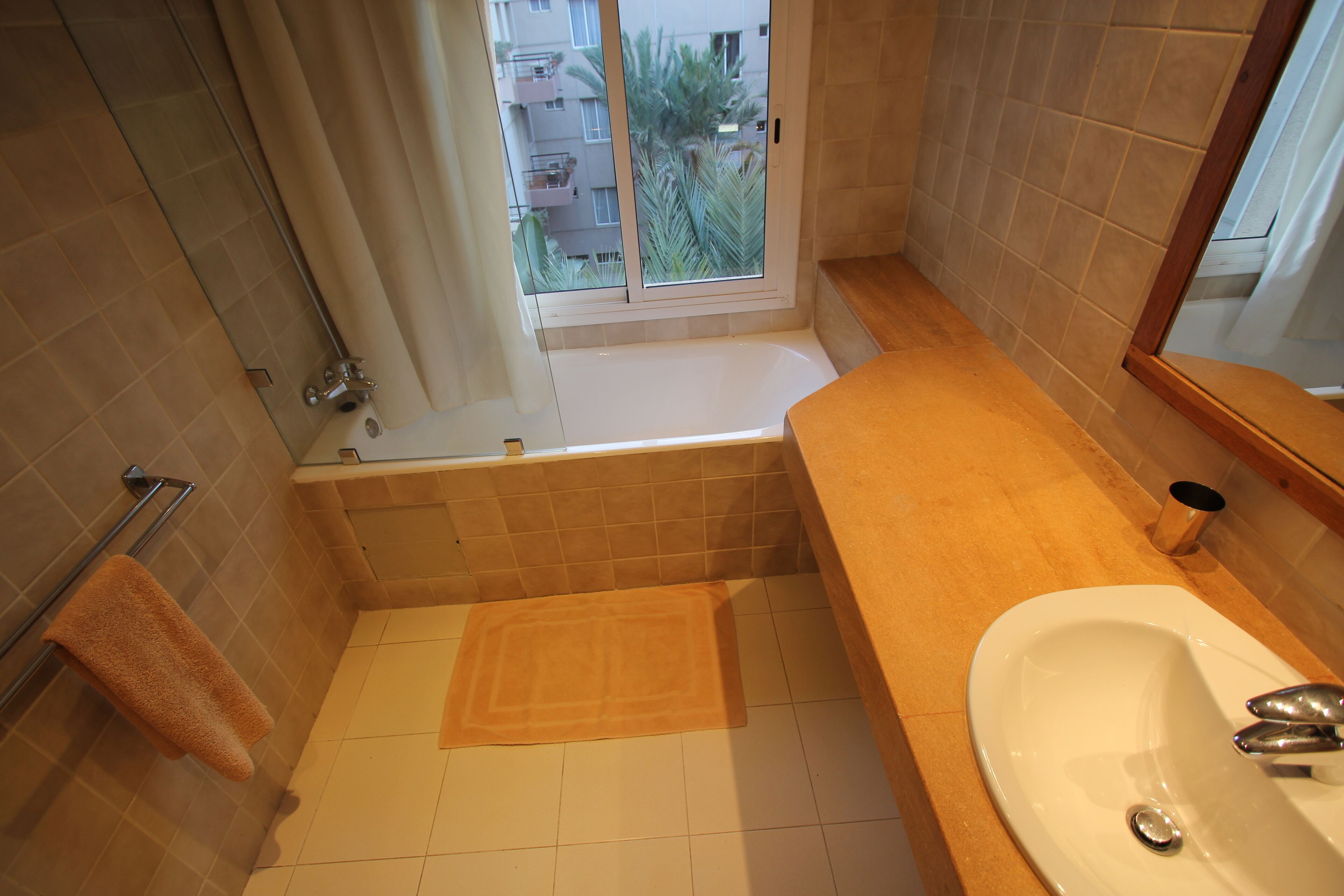 Une salle de bain avec une baignoire, un lavabo et une fenêtre donnant sur des immeubles et des palmiers.