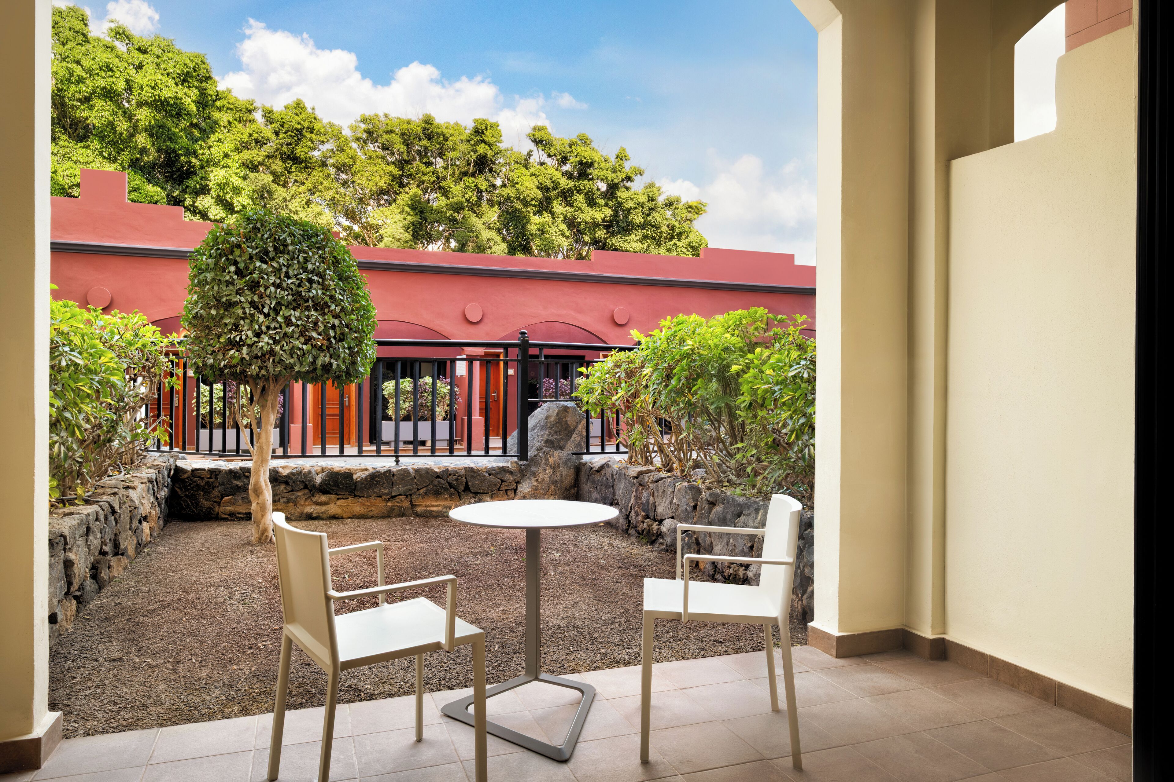Un patio con una mesa redonda y dos sillas, rodeado por un muro de piedra y vegetación.