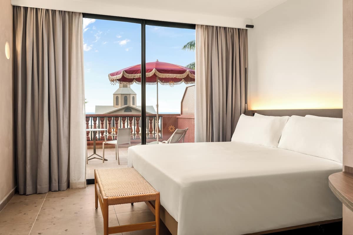 Ein Hotelzimmer mit einem großen Bett, einem kleinen Tisch und Blick auf ein Gebäude und eine Palme durch das Fenster.