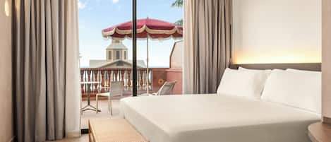 A hotel room with a large bed, a small table, and a view of a building and palm tree through a window.