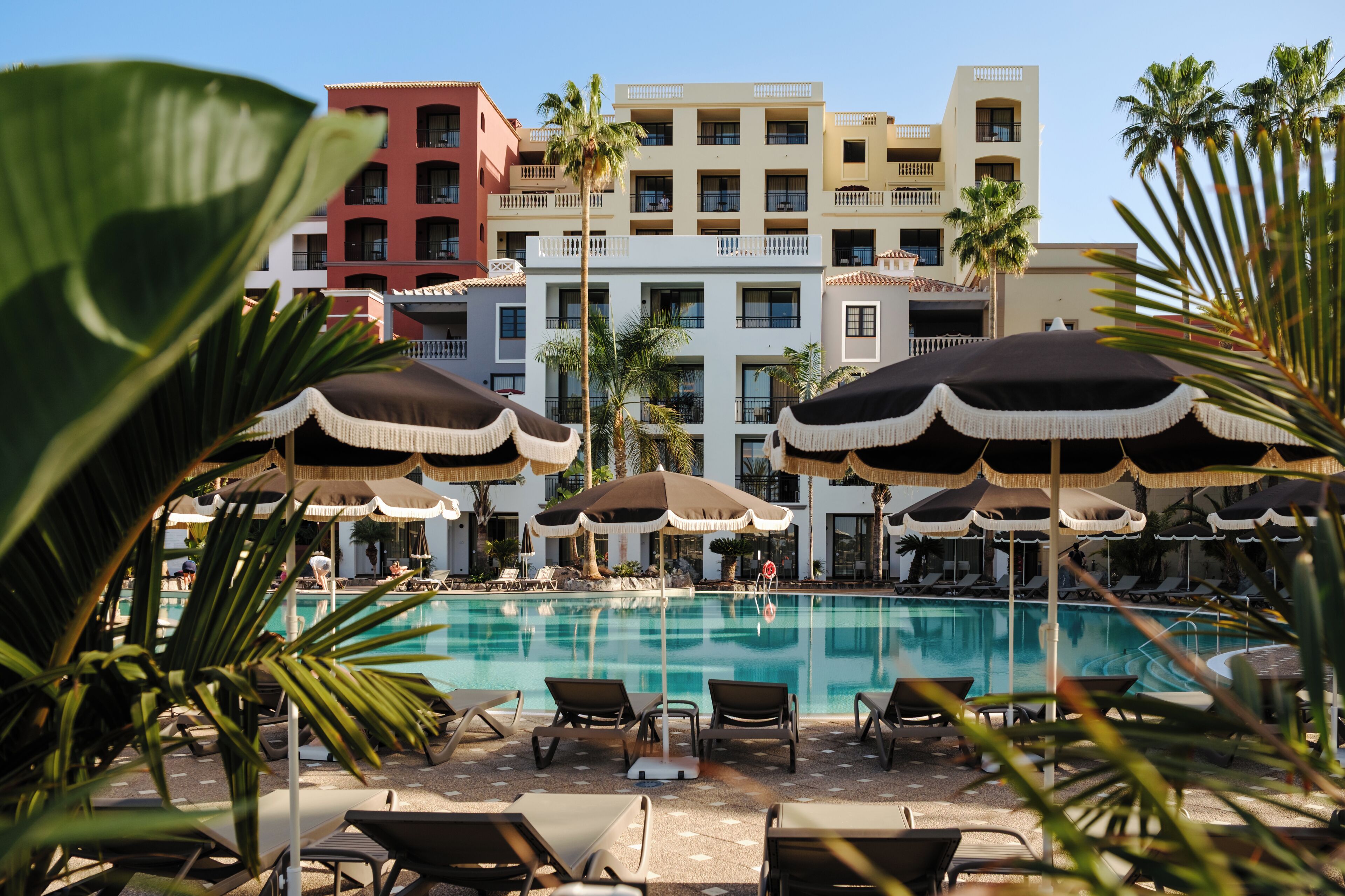 A pool area with lounge chairs and umbrellas, surrounded by palm trees and a multi-story building with balconies.