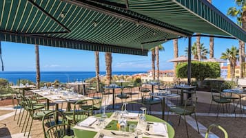 An outdoor dining area with tables and chairs, a covered patio, and a view of the ocean.