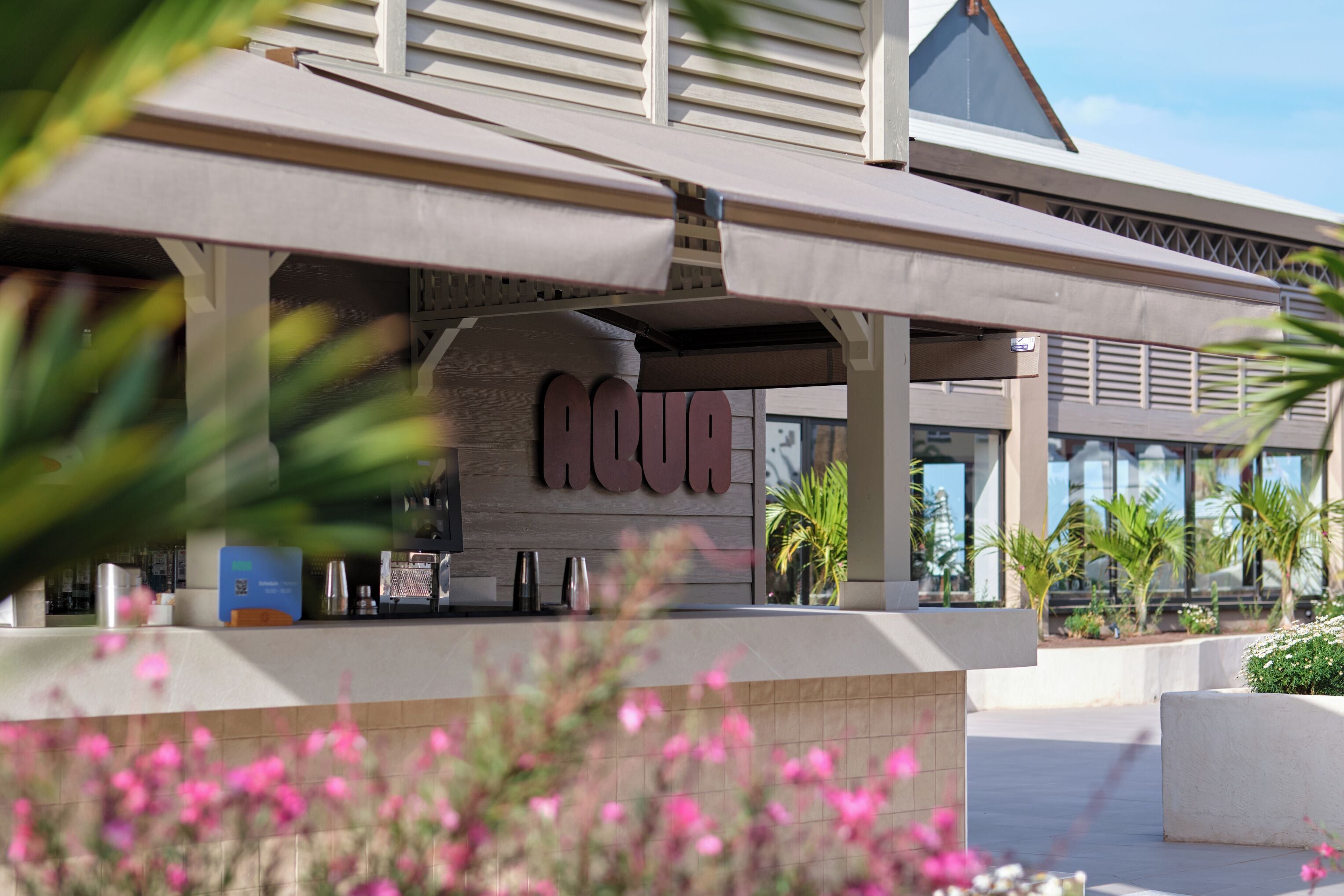 Entrada de un hotel con un llamativo letrero 'AQUA', zona de estar al aire libre y plantas en macetas.