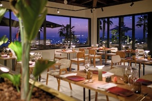 Restaurante com mesas preparadas para refeições, amplas janelas com vista e plantas dentro.