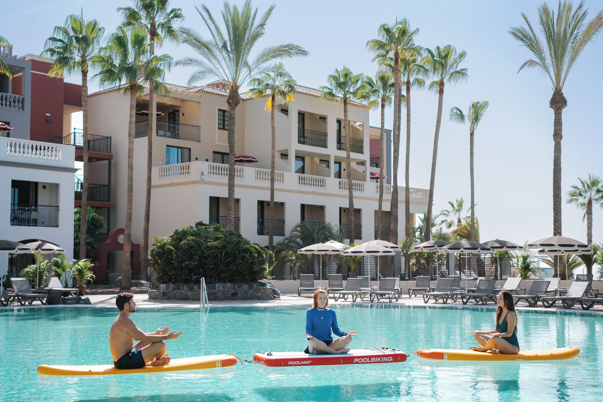 A pool area with three people meditating on surfboards, surrounded by palm trees and a hotel building with balconies.