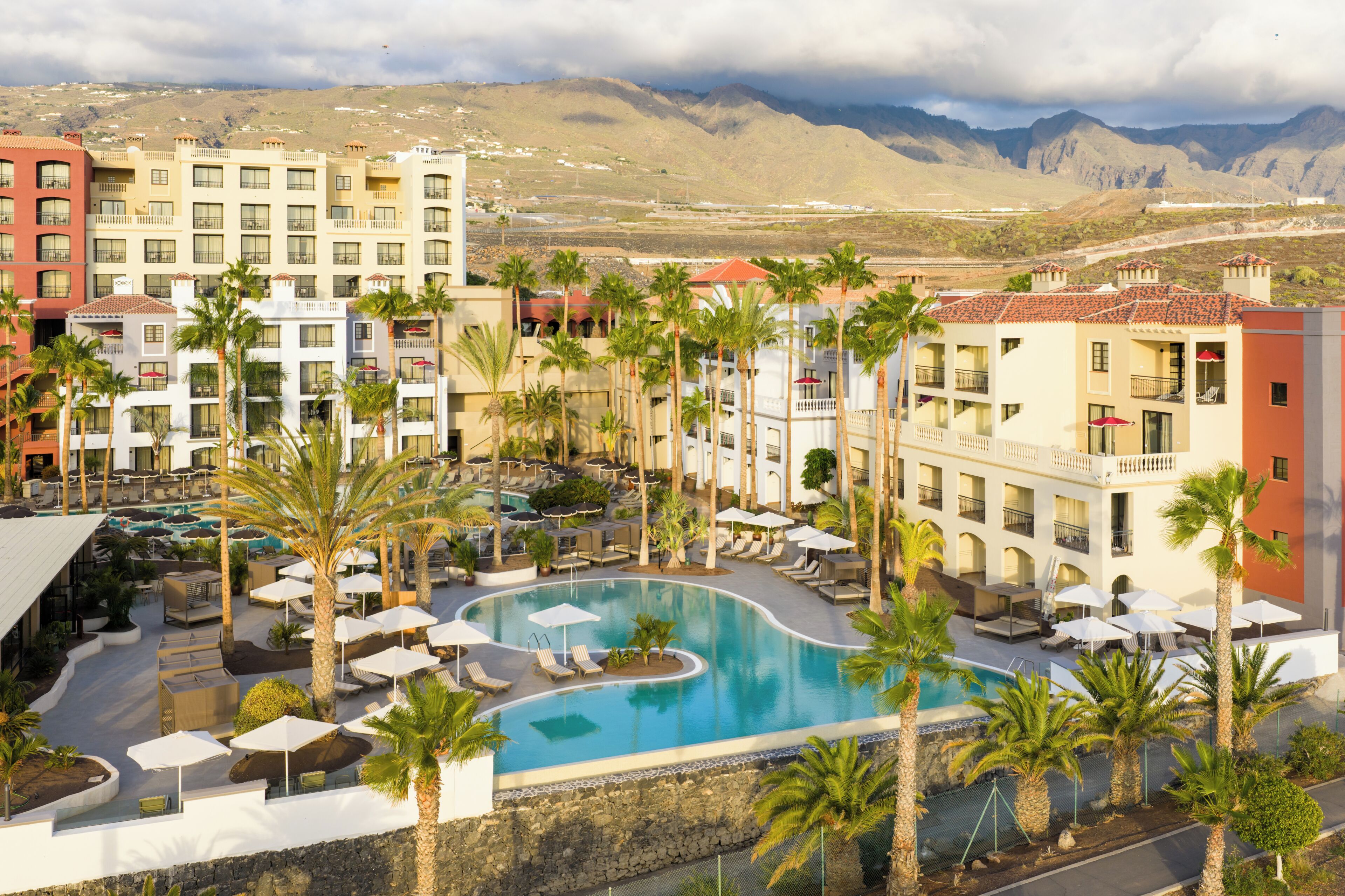 Un resort con piscina rodeada de palmeras y sillones de descanso, con montañas al fondo.