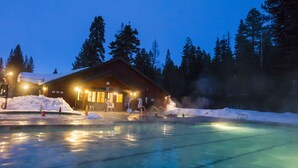 A heated outdoor swimming pool area at dusk with a wooden building and snow in the background.