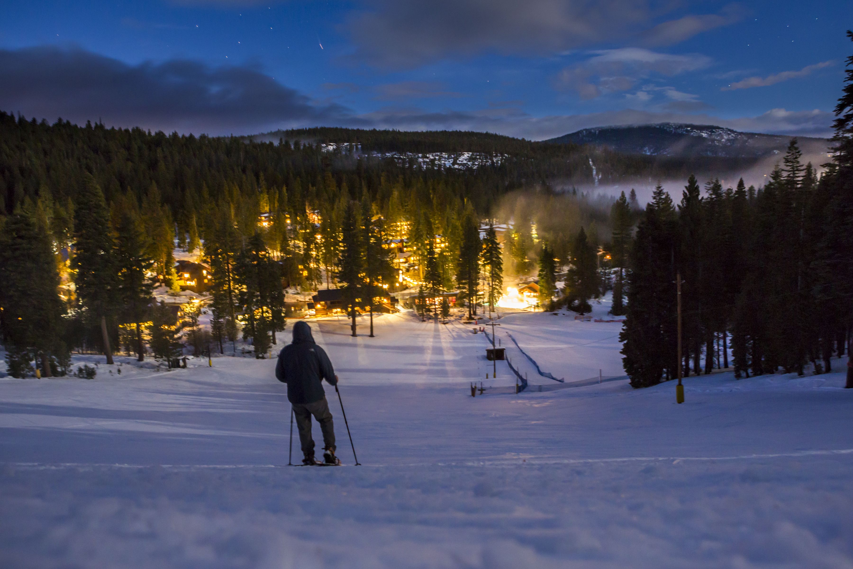 A person skiing on a snow-covered slope with a forested mountain in the background and a lit ski resort area.