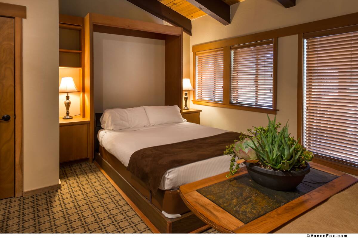 A bedroom with a bed, bedside lamps, a nightstand, a potted plant, and a window with blinds.