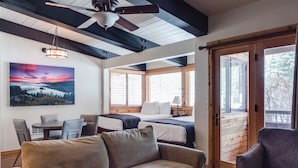 A cozy room with a sofa, a bed, a desk, and a chair. There is a window with wooden blinds and a door with a glass panel.