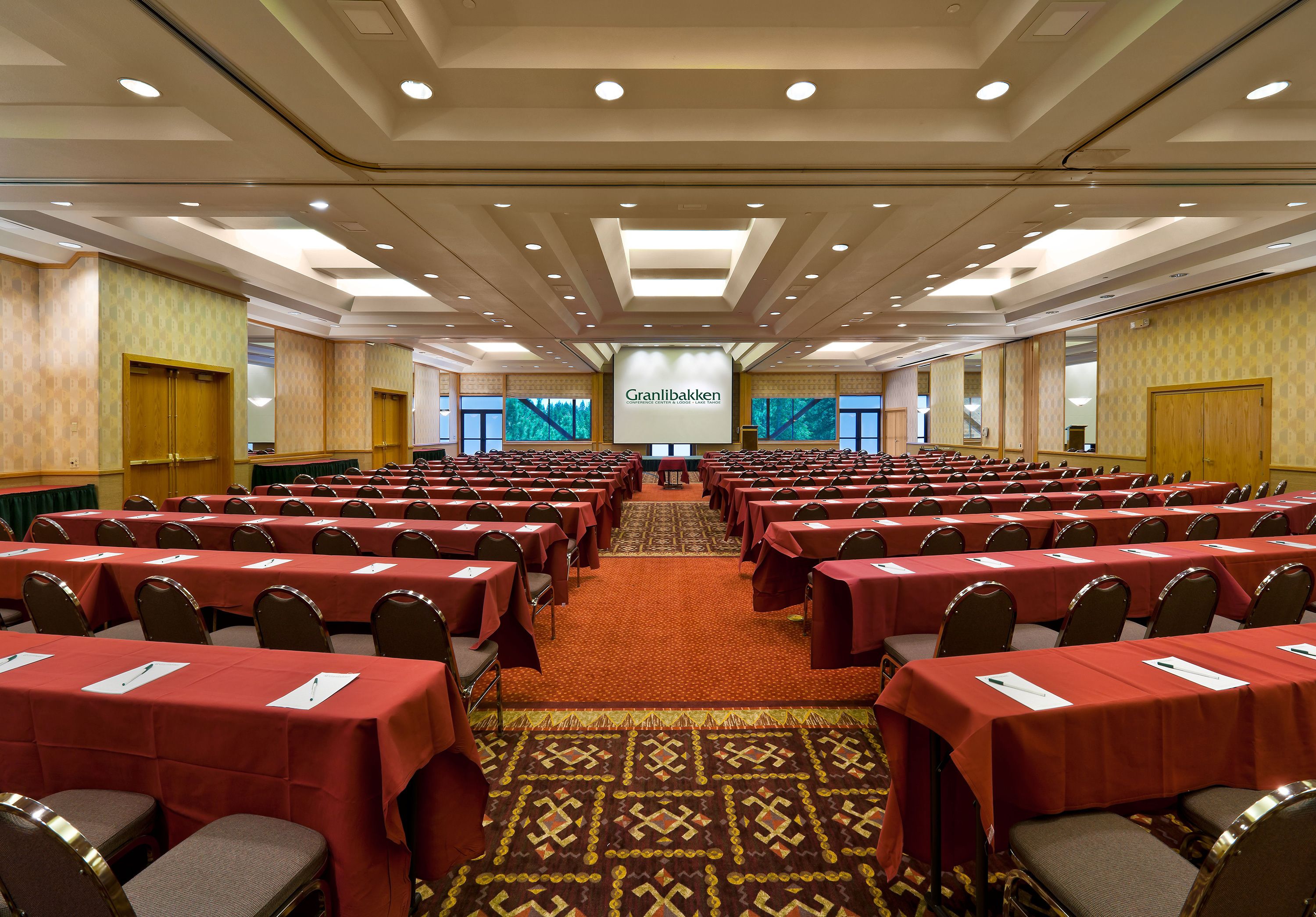 Une salle de conférence avec des rangées de tables et de chaises, une scène dotée d’un écran de présentation et un sol recouvert de moquette.