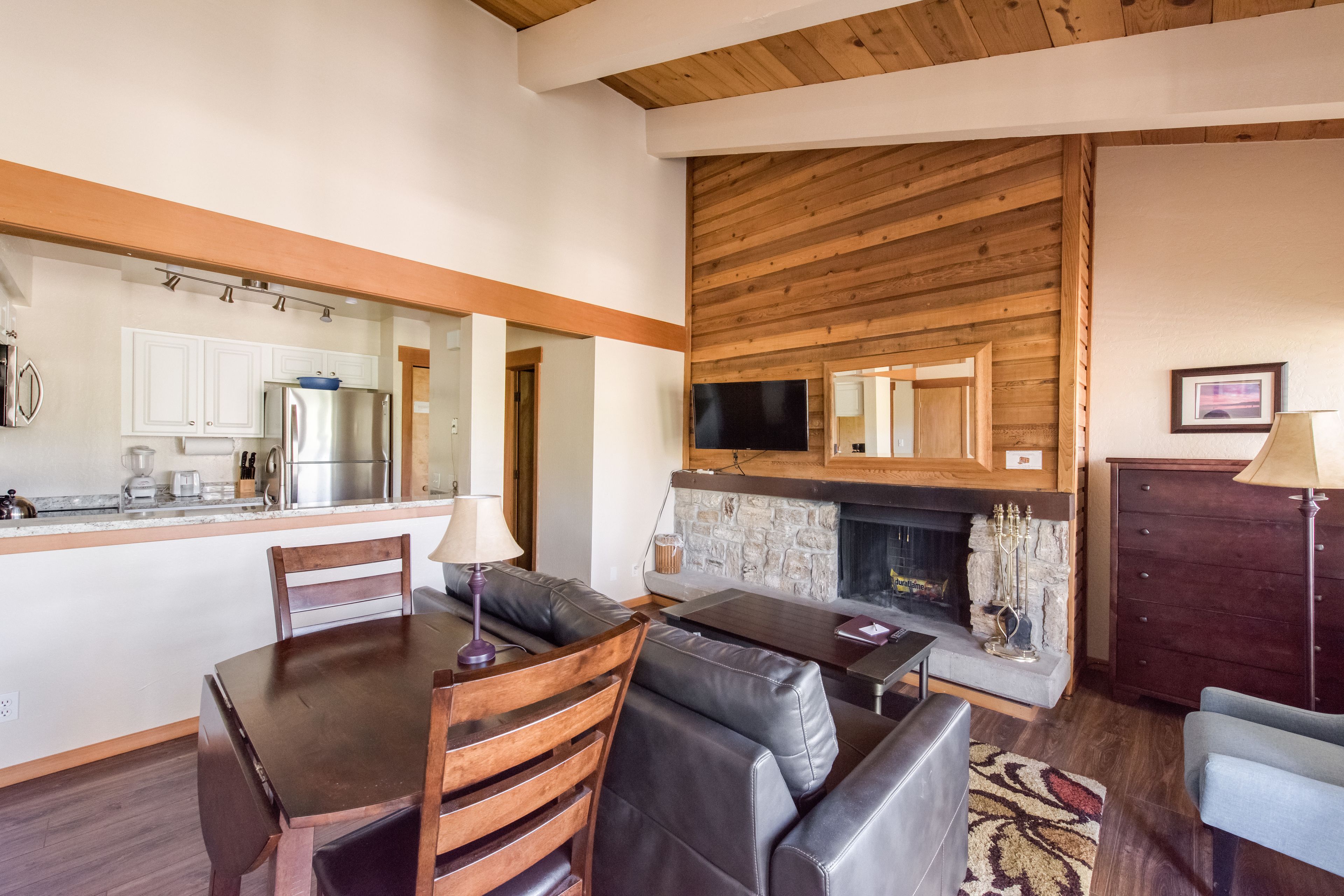 A modern kitchen with white cabinets, a stainless steel refrigerator, and a built-in oven. There is a dining area with a wooden table and chairs, and a living room with a fireplace and a television.