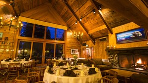 A cozy, wood-paneled dining area with tables set for a meal, a fireplace, and a large window offering a view of the night sky.