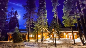 A lodge with a snow-covered landscape, surrounded by tall pine trees, and illuminated by warm interior lighting.