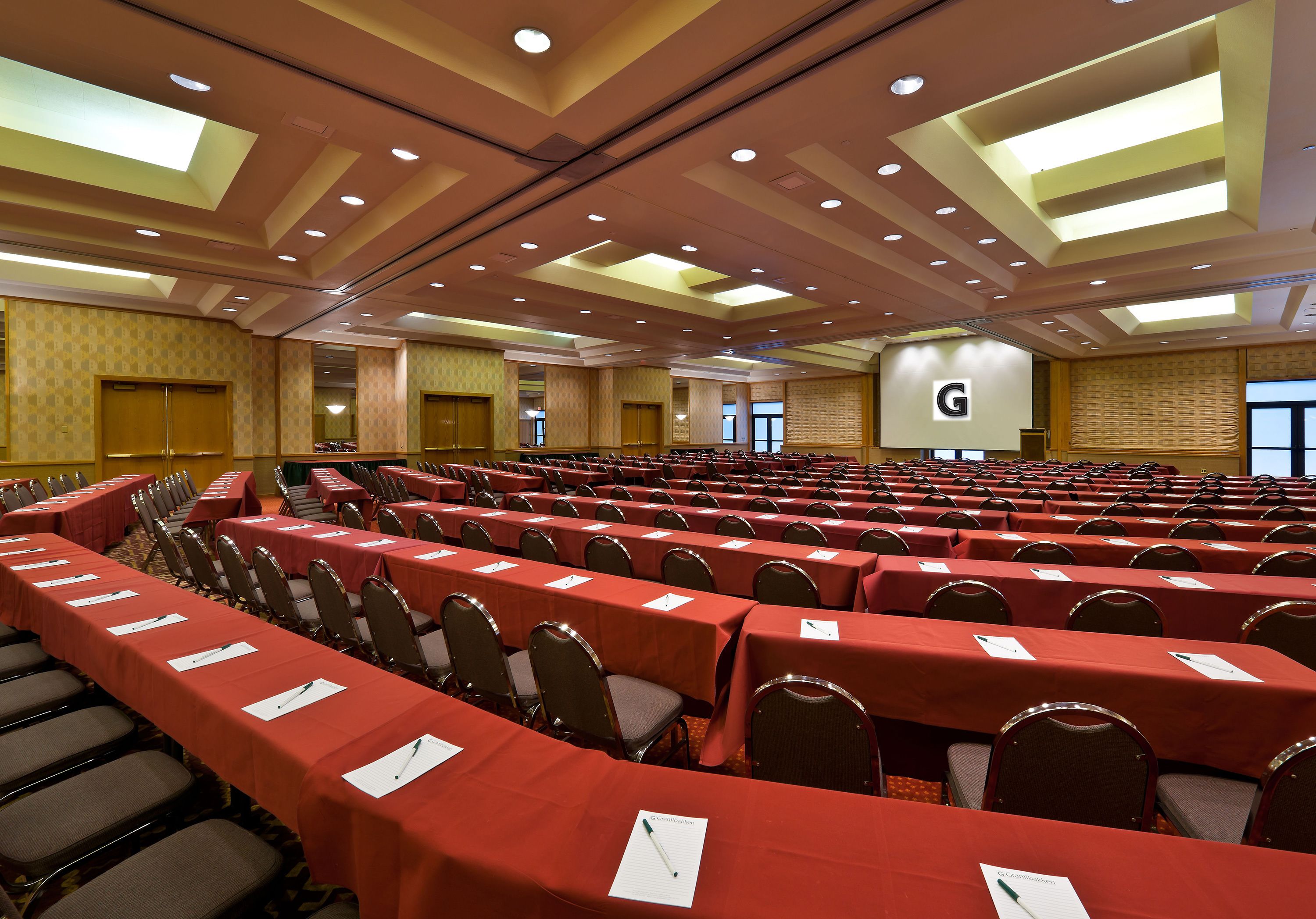 A conference room with rows of tables and chairs, a large screen, and a 'G' logo on the wall.