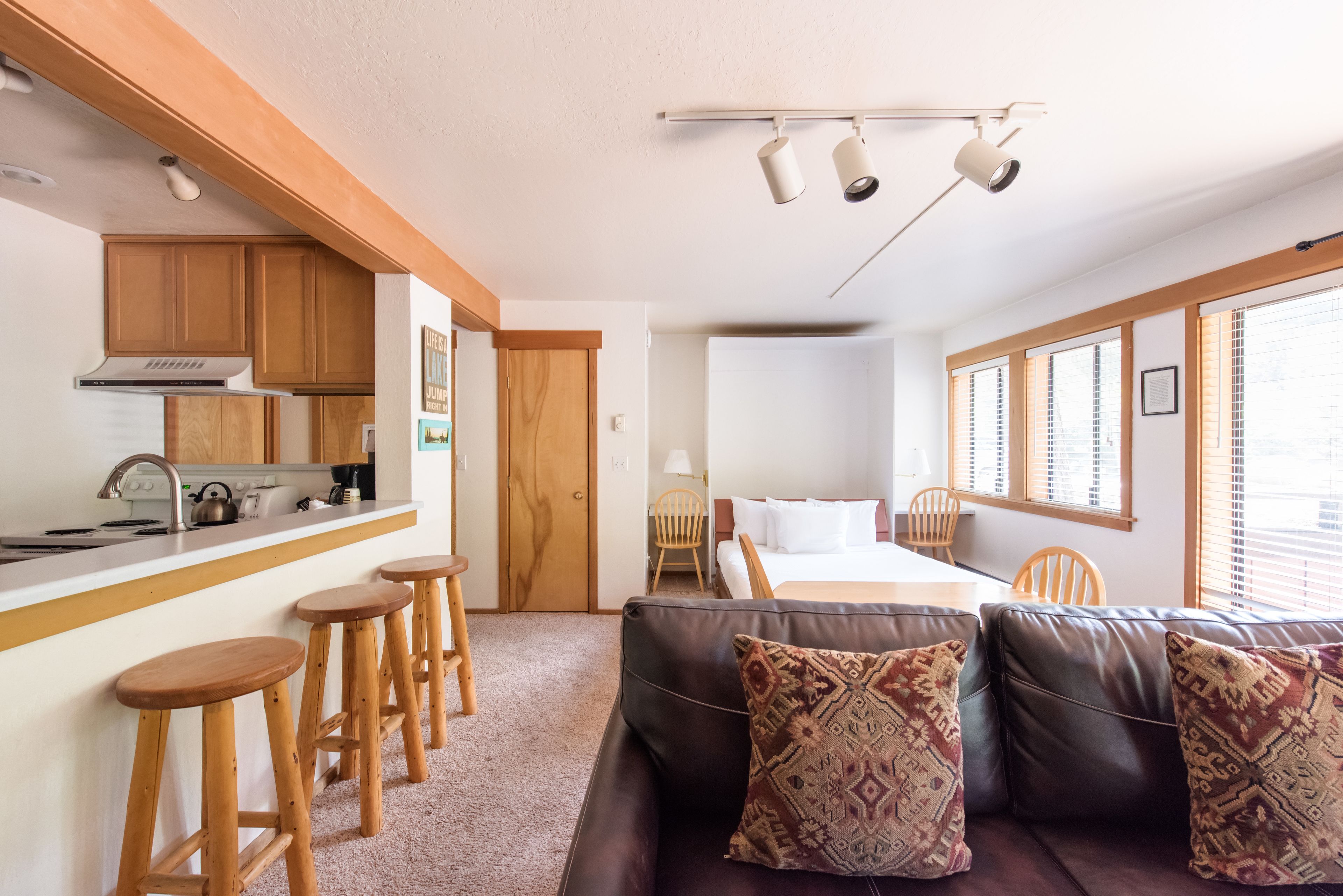 A compact, modern room with a kitchenette, a sofa with patterned cushions, and a bed with a wooden headboard.