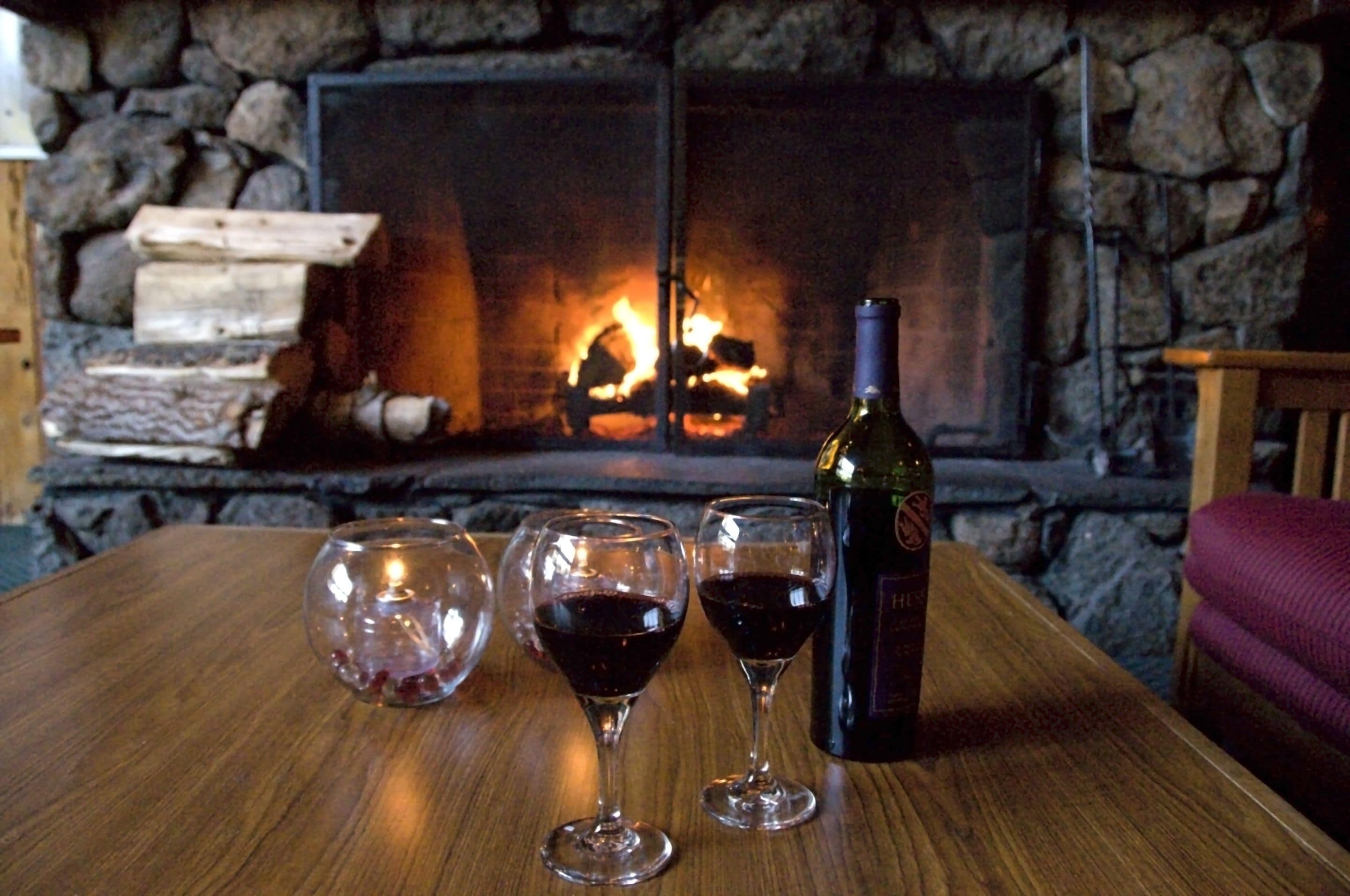 Une table en bois, avec trois verres à vin et une bouteille de vin, placée devant une cheminée en pierre où brûle un feu.