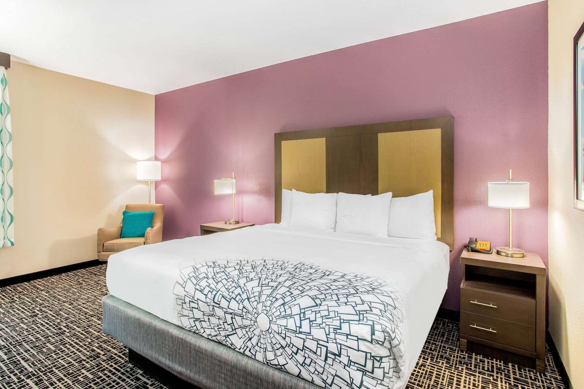 Habitación de hotel con una cama grande, una silla, dos mesitas de noche con lámparas y una alfombra estampada.