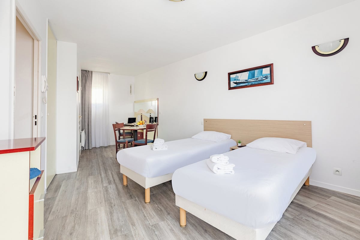 A hotel room with two single beds, a desk with a chair, a small table with a lamp, and a painting on the wall.