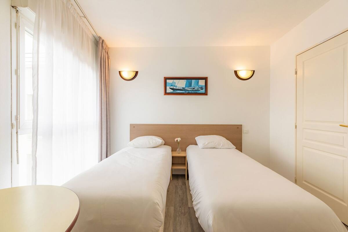 A hotel room with two single beds, a small table, a painting on the wall, and two wall-mounted lights.