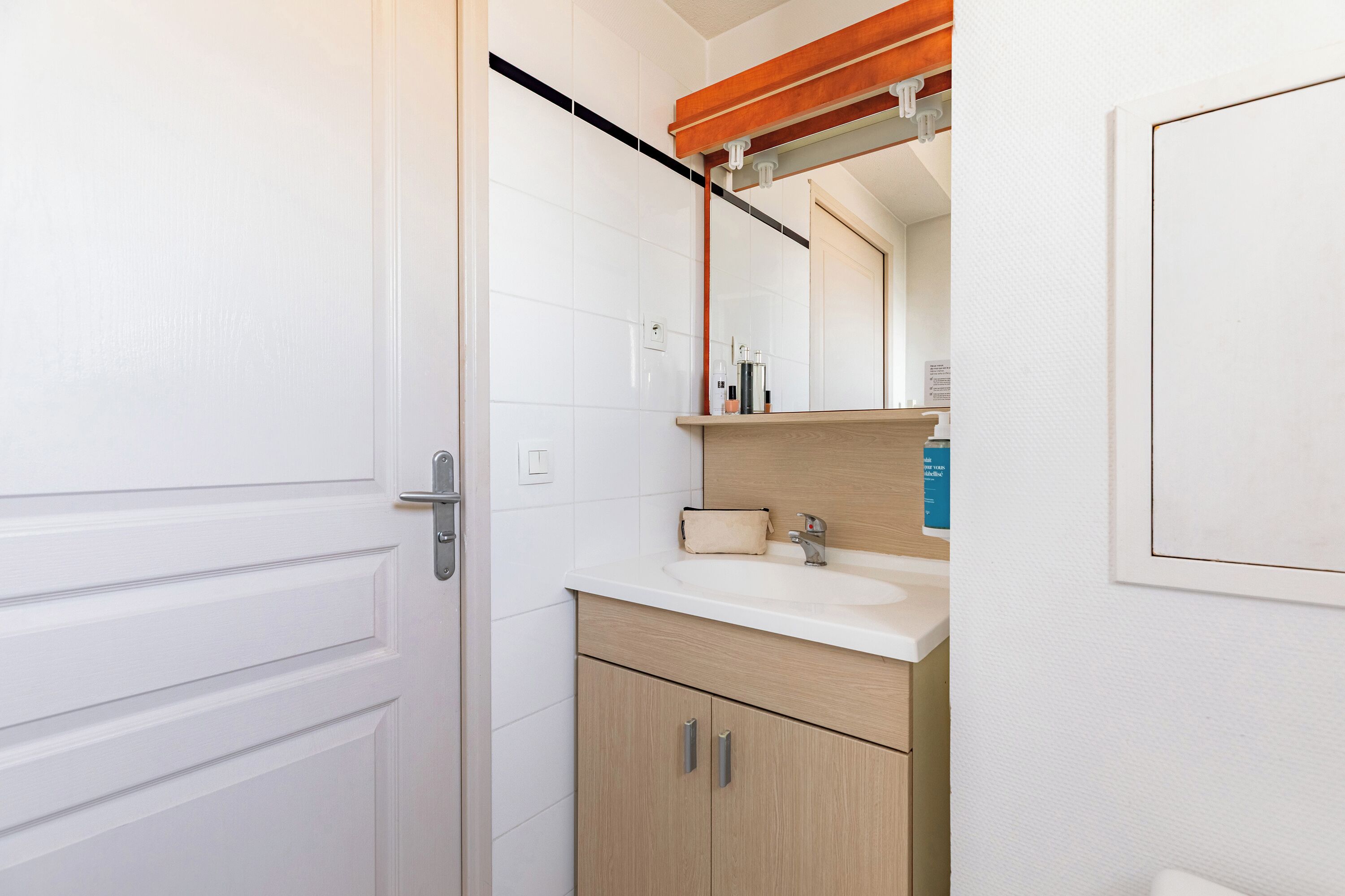 Un baño con una puerta blanca, un tocador de madera con un lavabo y un espejo sobre el tocador.