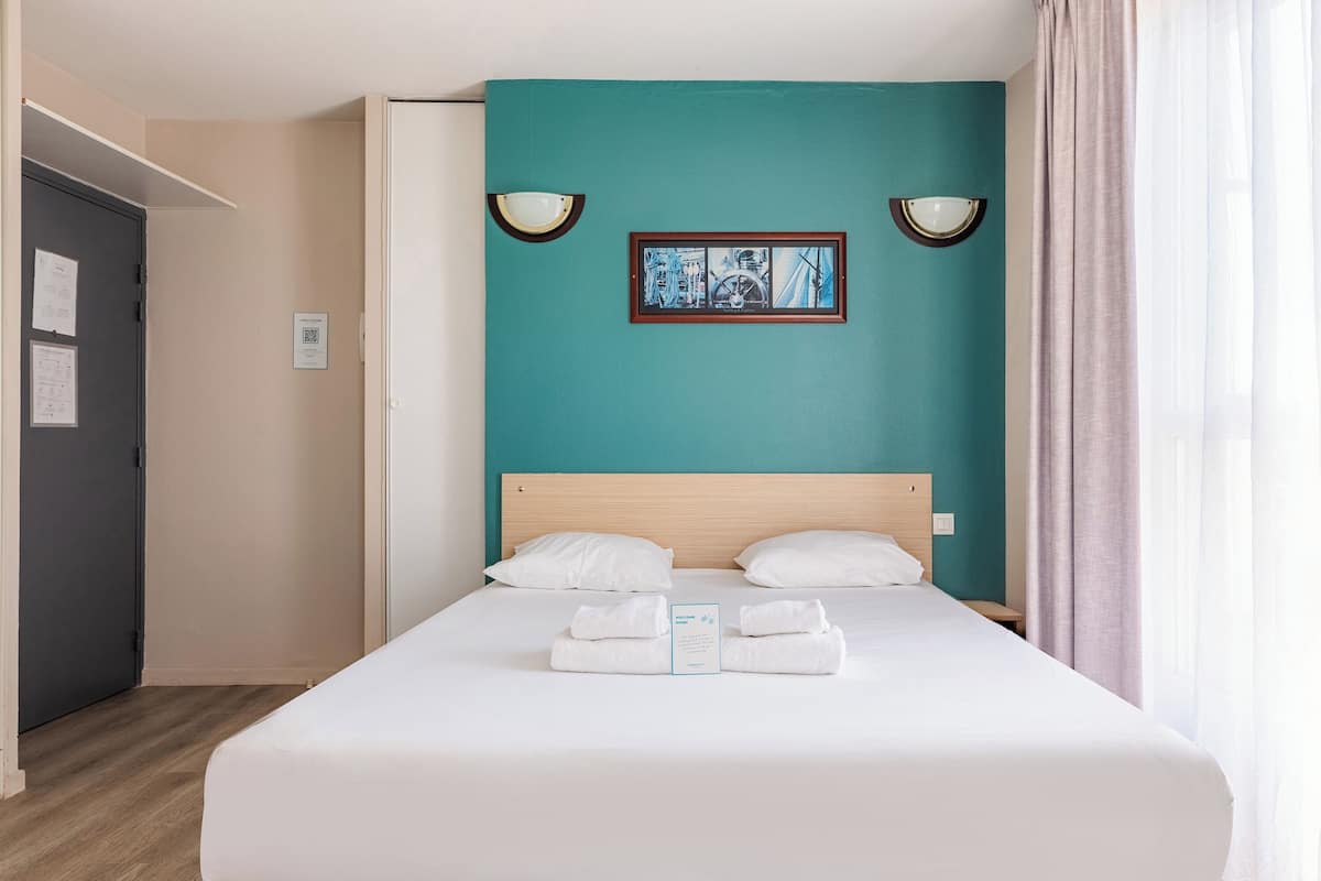 A neatly made bed with white linens and towels, a teal accent wall, and a framed artwork above it.